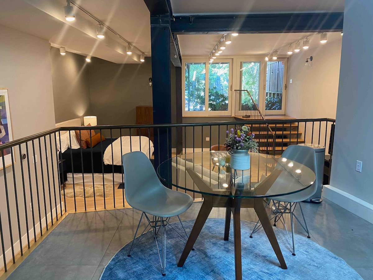 A modern dining area is highlighted by a glass table and mid-century style chairs. Soft lighting illuminates the space, which features a glimpse of a cozy sleeping area with a bed in the background. Large windows invite natural light, enhancing the open feel.