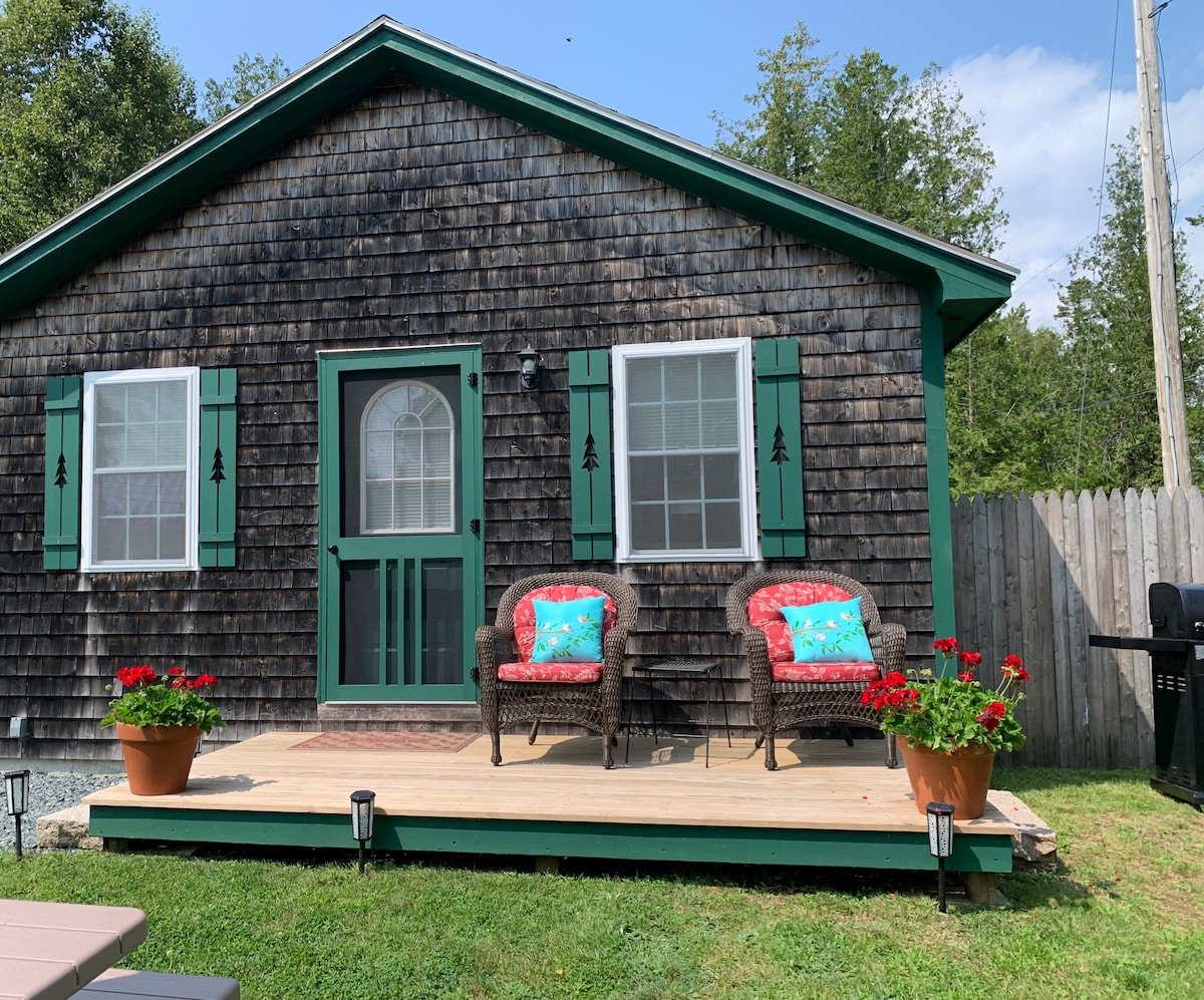 The cottage's front exterior features a wooden facade, complemented by green shutters and two inviting chairs adorned with colorful cushions on the porch. Potted flowers add a touch of vibrancy to the outdoor space, enhancing the overall welcoming appearance.