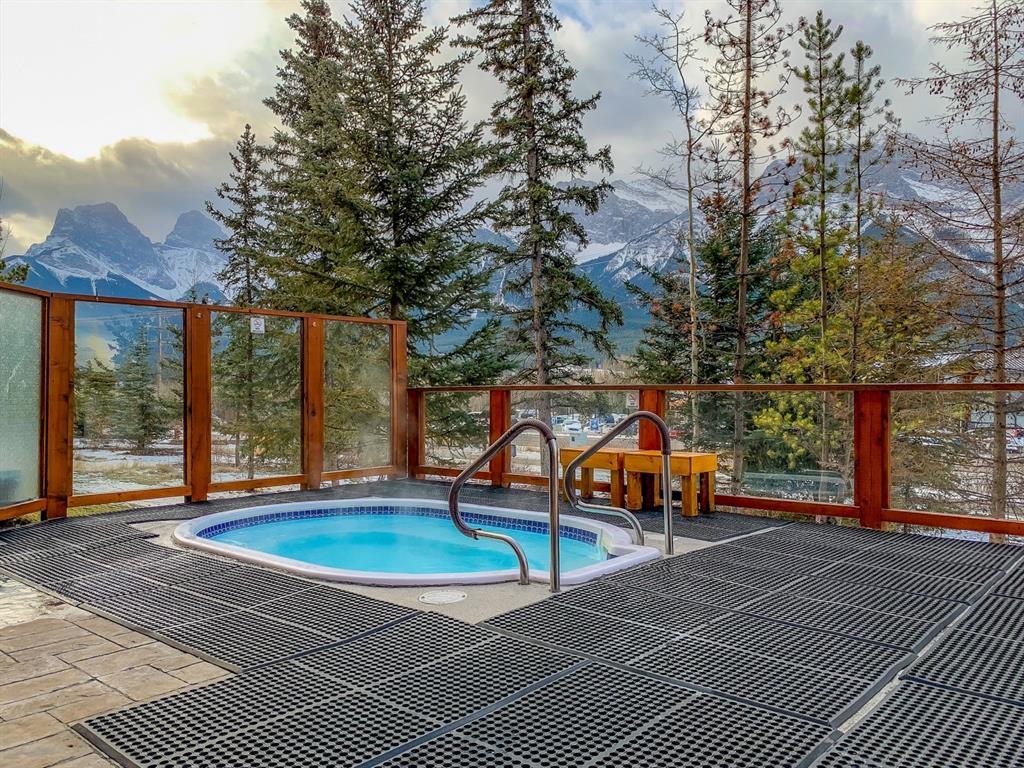 A hot tub is situated on a deck surrounded by trees, offering a secluded atmosphere. The surface is lined with textured flooring for safety. Views of distant mountains can be seen in the background, with a soft light illuminating the scene.