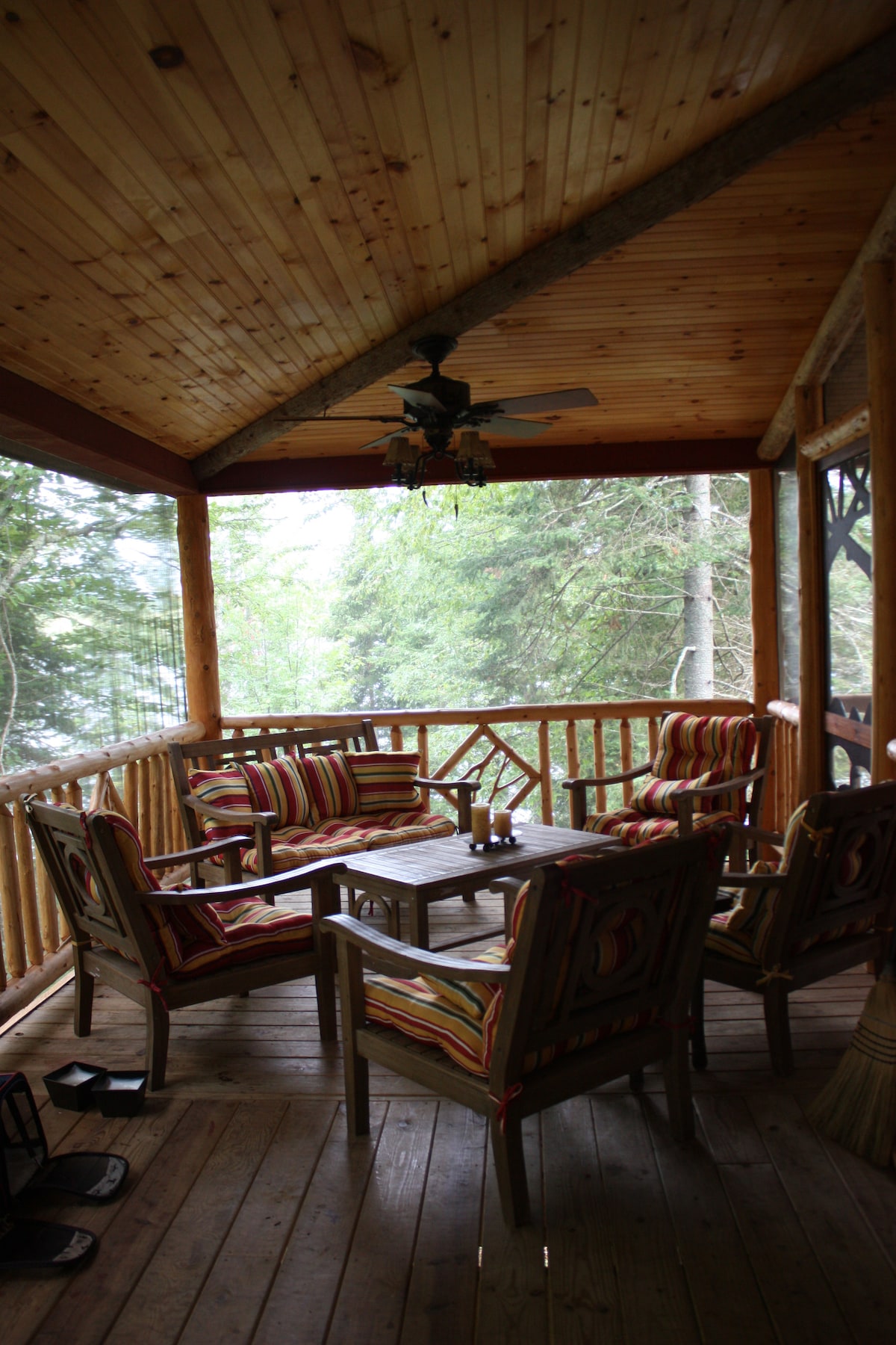 A spacious screened porch is furnished with comfortable seating, featuring patterned cushions. A wooden table is centered among the chairs, creating a cozy gathering space. Natural light filters through the screens, and views of the surrounding trees are visible.