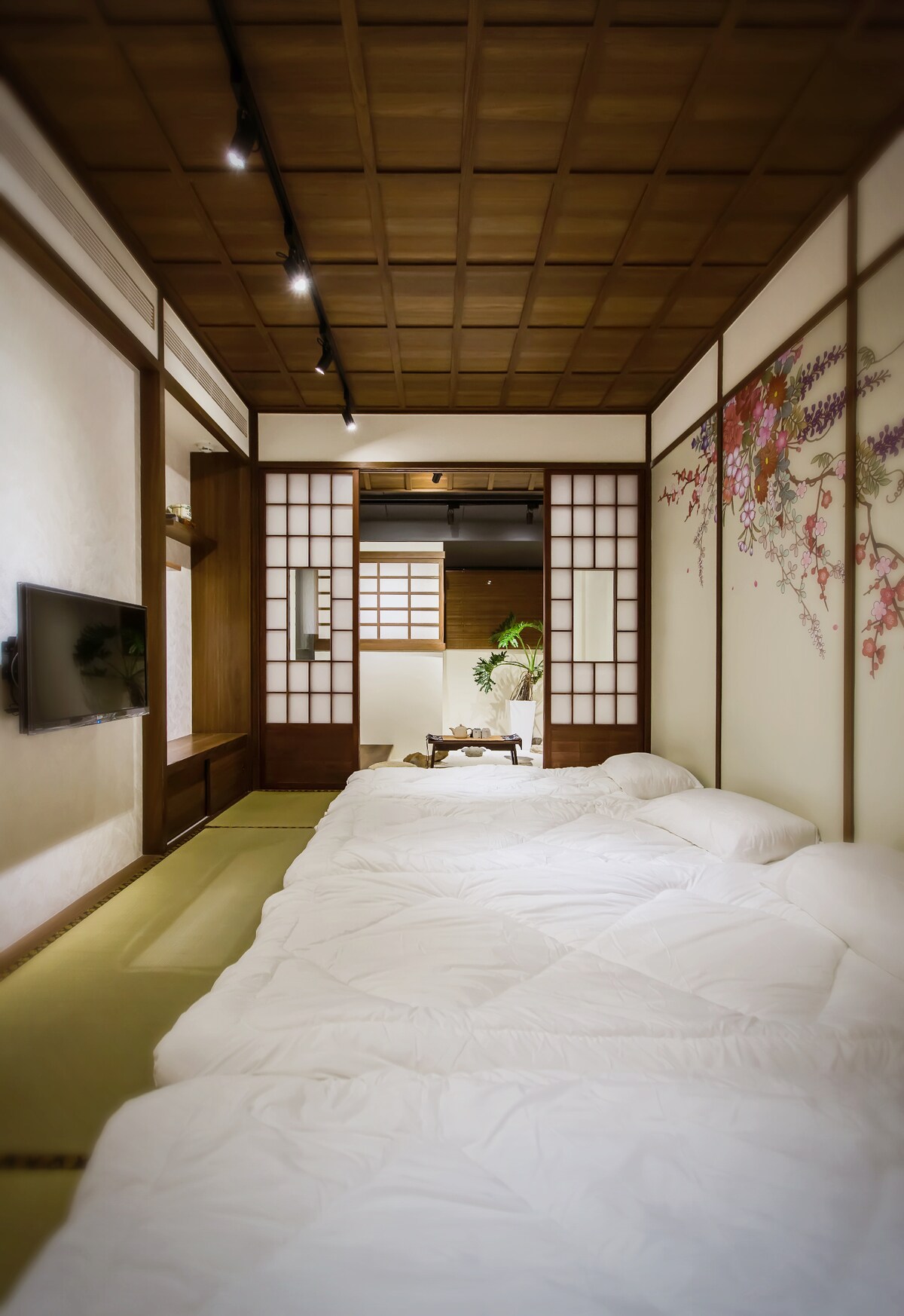 A serene sleeping area features traditional tatami mats and spacious bedding arranged neatly on the floor. Soft, neutral tones are enhanced by wooden beams and a floral mural on one wall. A television is mounted for entertainment, while a glimpse of a green plant adds a touch of nature.