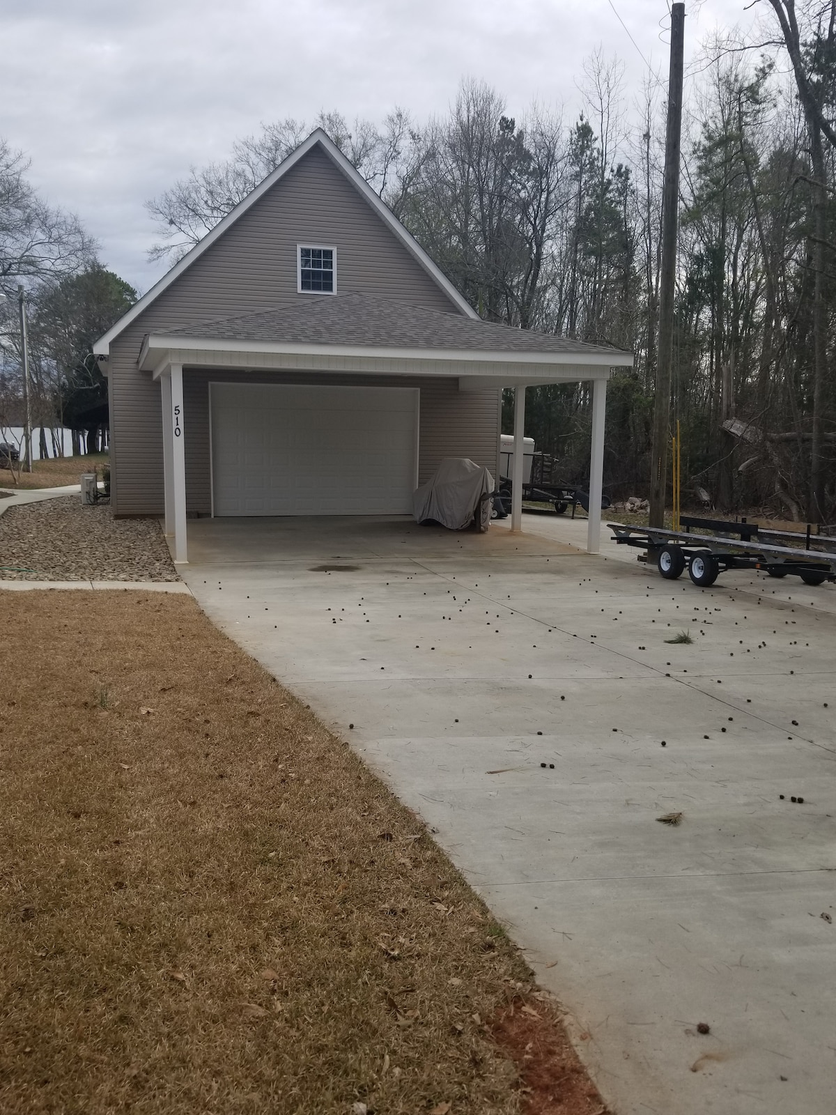 A driveway leads to a house with a covered front porch and a garage. Gravel is present on one side of the pavement. Tall trees line the background, contributing to a natural setting. A trailer is parked beside the driveway.