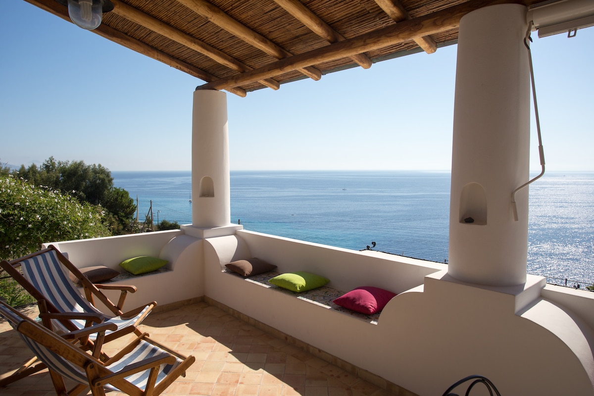 A spacious terrace is framed by two white columns with charming stone accents, offering a breathtaking view of the sea. Comfortable cushions in various colors are placed on the built-in seating, while a wooden lounge chair invites relaxation. Sunlight brightens the serene space.