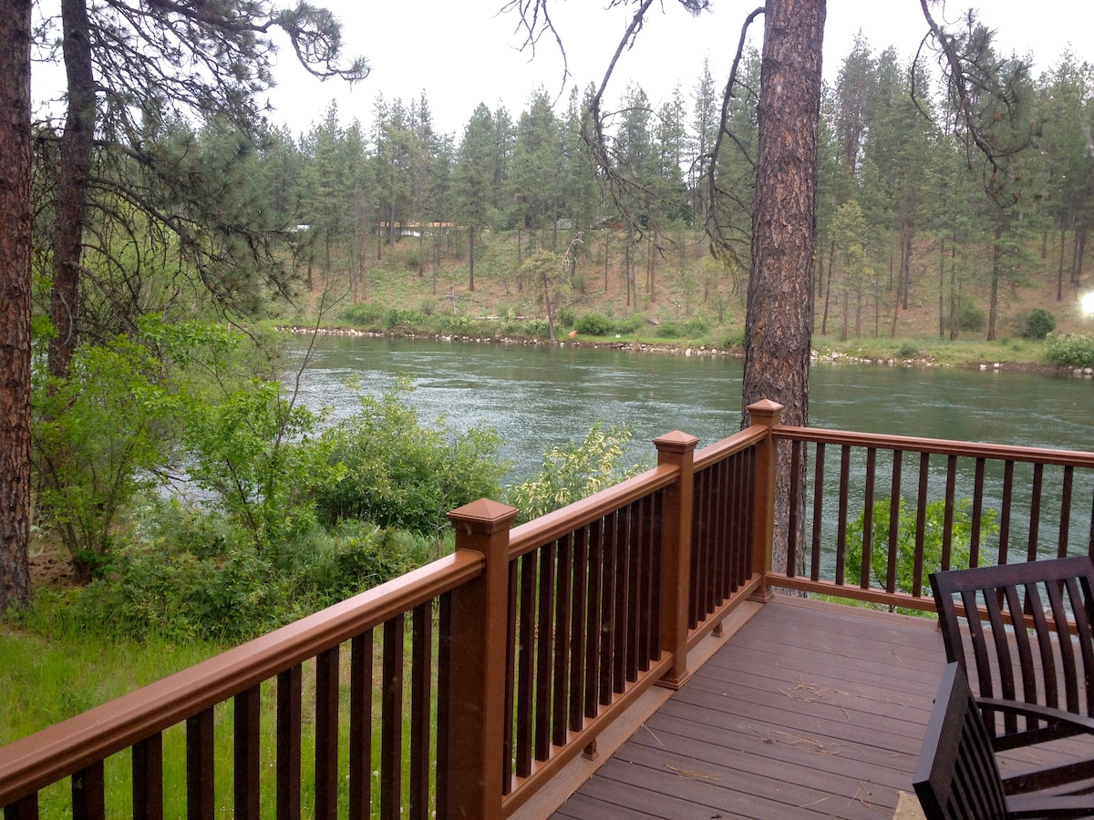A serene view from the wooden deck overlooks the flowing Spokane River. Lush greenery and trees line the banks, creating a peaceful and natural setting. The wood railing adds a rustic touch, enhancing the connection with the surrounding landscape.