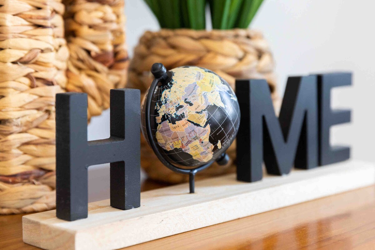A decorative tabletop display features the word 'HOME' spelled out in bold black letters, positioned beside a small globe. The globe, showcasing the continents in muted colors, is supported by a simple black stand. Natural woven baskets and green plants are visible in the background.