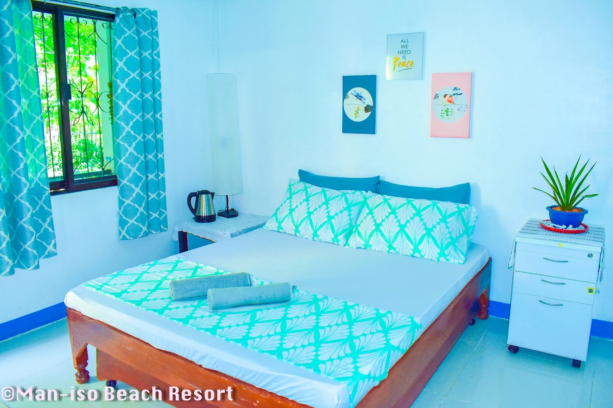 A well-lit room features a spacious bed adorned with a patterned turquoise and white bedspread. Nearby, a small table holds a kettle while a cabinet provides storage. Two framed artworks and a potted plant add color to the serene, white-walled space.