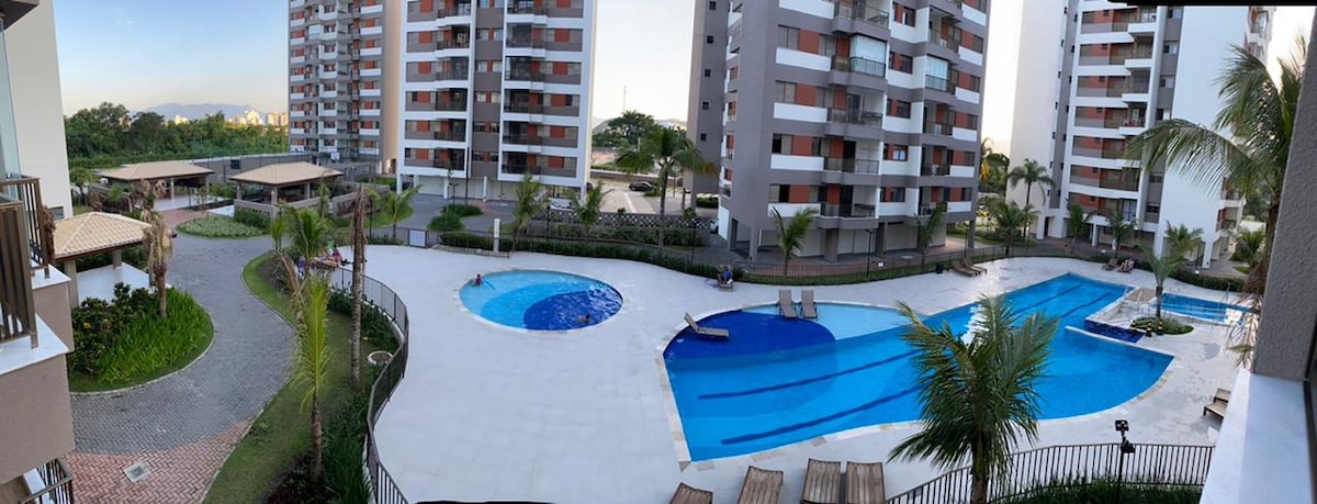 A serene outdoor area features two swimming pools surrounded by well-maintained landscaping. Lounge chairs are positioned near the water's edge, providing space for relaxation. The nearby buildings have modern designs with balconies overlooking the pools, enhancing the tranquil environment.