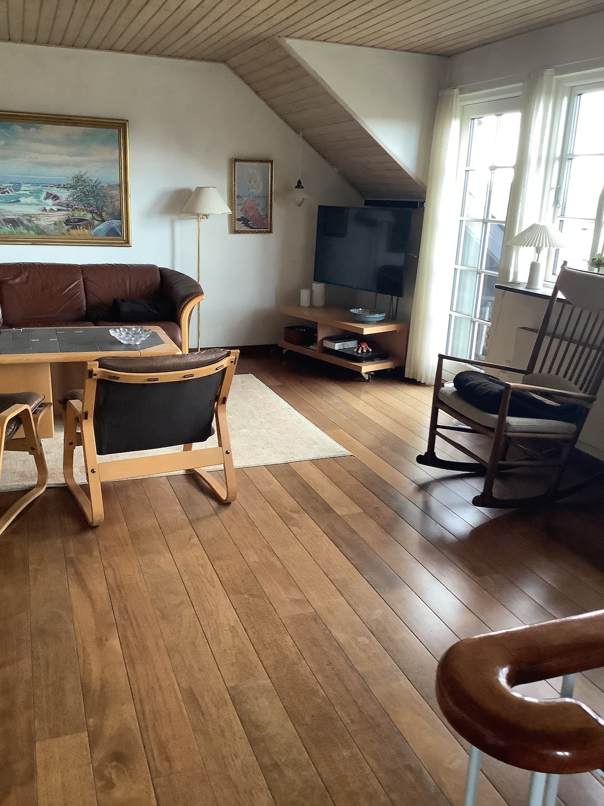 A comfortable living space is furnished with a brown leather sofa and a wooden rocking chair. A coffee table sits atop a light area rug, while a TV is positioned on a low wooden stand. Natural light floods in through large windows, illuminating the wooden floor.
