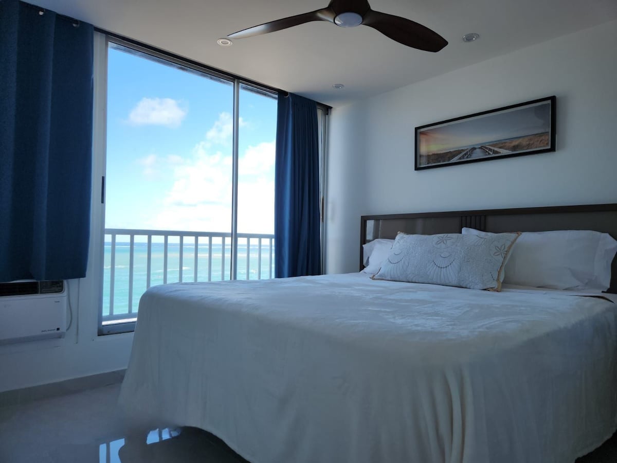 A spacious bedroom features a large bed with soft linens, complemented by a decorative pillow. Floor-to-ceiling windows provide views of the ocean and allow natural light to fill the room. A ceiling fan and air conditioning unit are also visible.