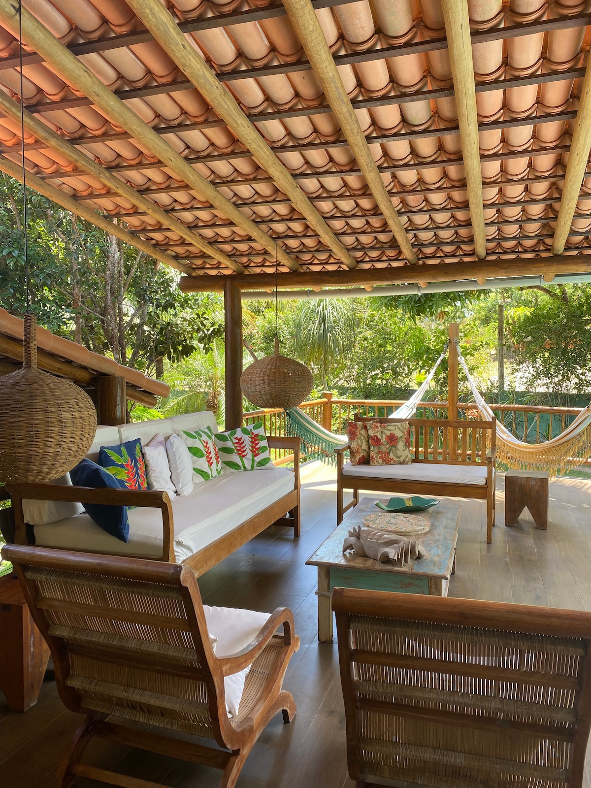 A covered outdoor lounge area features a wooden sofa adorned with colorful cushions, accompanied by two woven chairs. A rustic coffee table sits in the center, while a hammock is suspended nearby. Lush greenery surrounds the space, enhancing the natural atmosphere.