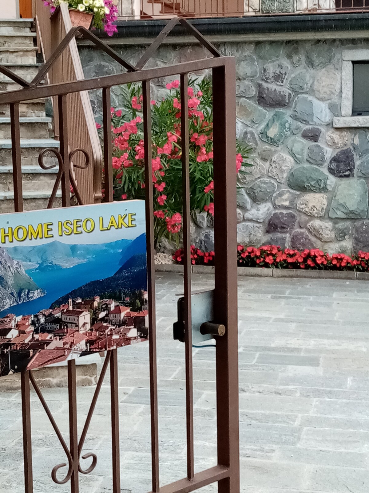 The image features a metal gate with a sign reading 'HOME ISEO LAKE.' In the background, stone steps lead to a textured stone wall, while vibrant pink flowers bloom near the entrance. A neatly paved surface is visible, contributing to a welcoming outdoor space.