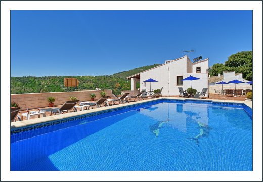 A shared swimming pool is featured, surrounded by sun loungers and shaded by blue umbrellas. The clear water reflects the bright blue sky and lush green hills in the background, offering a serene setting for relaxation and leisure.
