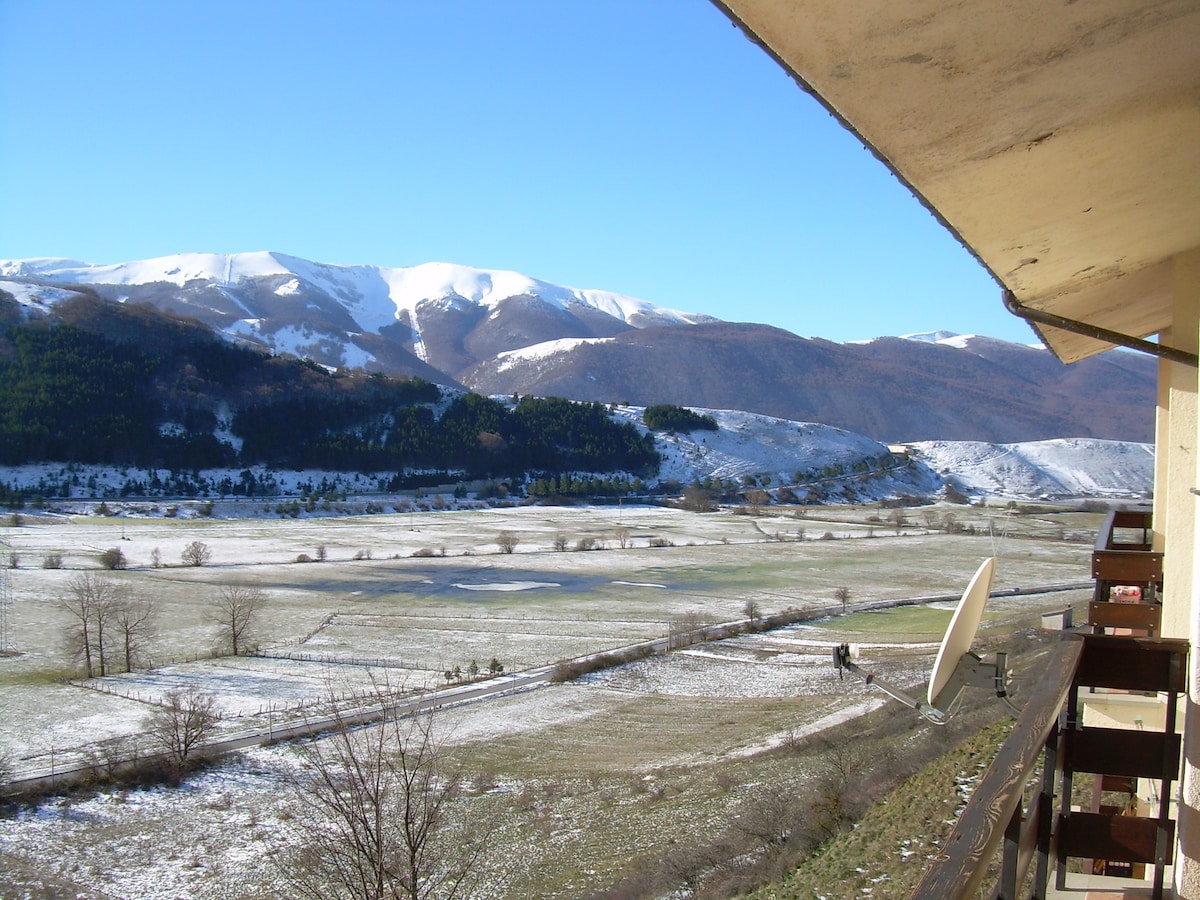 A breathtaking view of snow-capped mountains is visible from the balcony, overlooking a vast, open valley. The landscape features a mix of grassy areas and patches of white snow, while trees dot the distant hills under a clear blue sky.