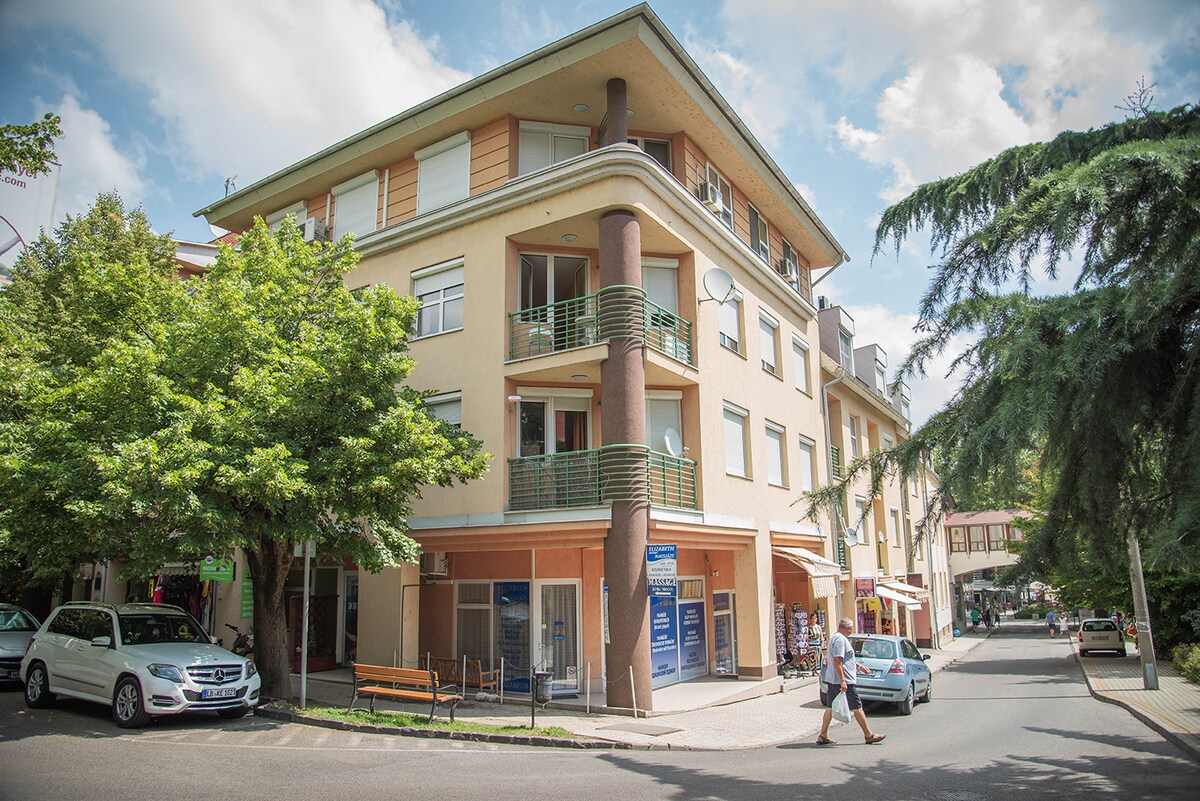 A modern three-story building is situated on a quiet street, featuring large balconies and a welcoming facade. Surrounding trees provide shade, and parked cars line the street. Various shops and eateries can be seen in the vicinity, enhancing the neighborhood's accessibility.