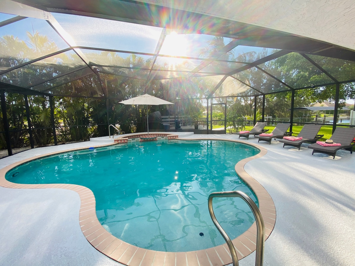 A spacious outdoor pool area features a heated pool surrounded by a sun deck. Lounge chairs are positioned for relaxation under a bright sky. An umbrella provides shade, enhancing comfort. Lush greenery frames the area, contributing to a serene atmosphere.