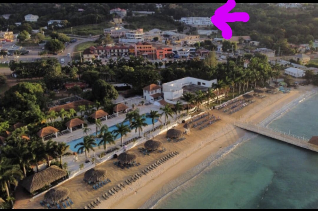 An aerial view captures a coastal scene with sandy beach, palm trees, and a clear water shoreline. A pool is visible near the beach, flanked by loungers. Nearby, numerous buildings and resorts can be seen, surrounded by lush greenery in the background.