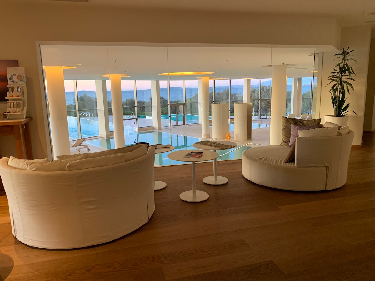 A modern sitting area features soft, round chairs paired with minimalist side tables. A large glass wall offers an unobstructed view of an indoor pool area, where natural light floods in, creating a serene ambiance. Lush greenery is visible in the background.