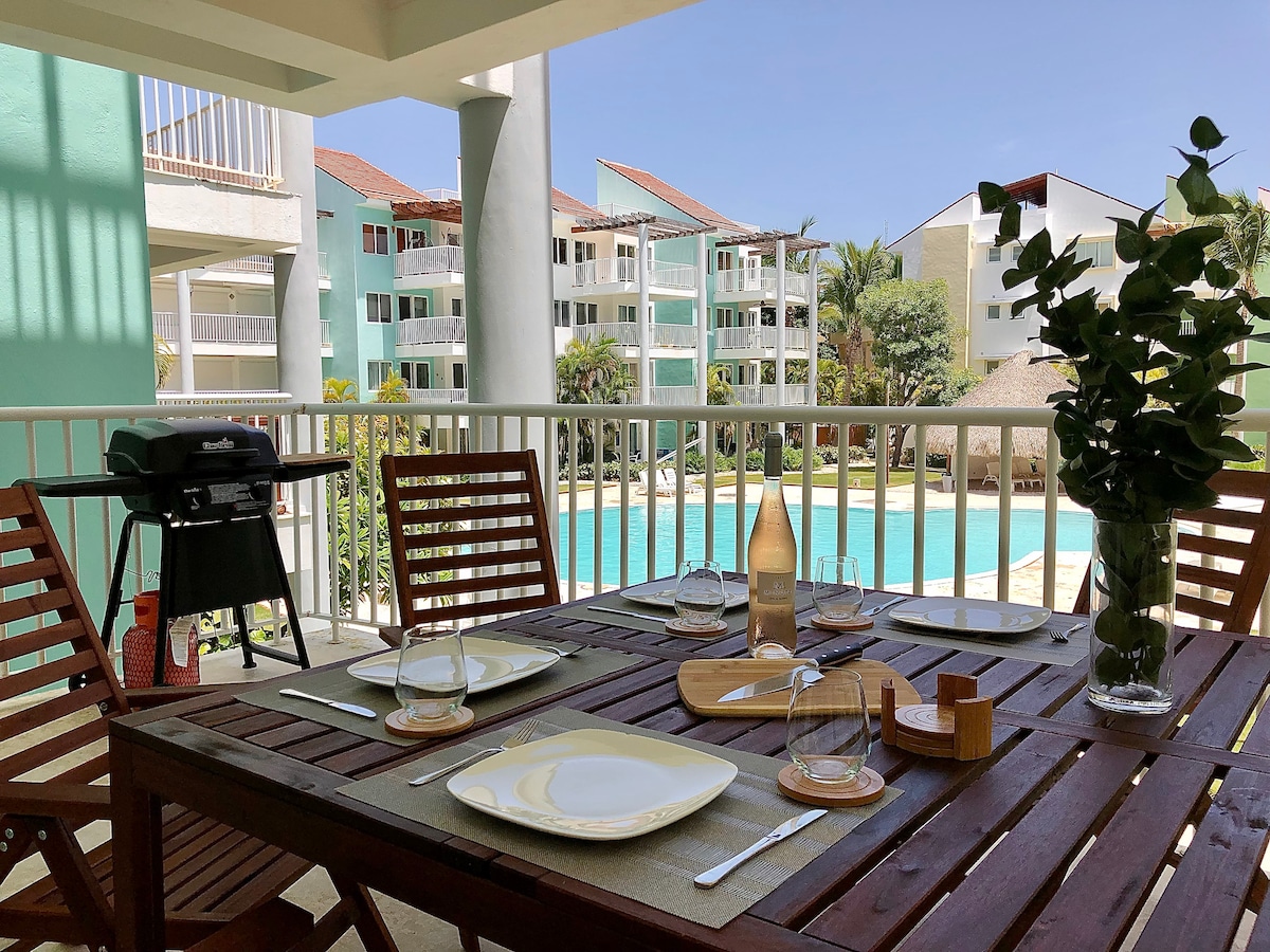A covered terrace features a wooden dining table set for six, complete with dishes and glasses. A charcoal grill sits nearby. Views of the landscaped gardens and a swimming pool are visible in the background, under a clear blue sky.