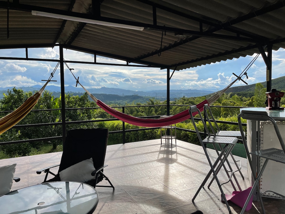 A covered terrace is showcased, featuring comfortably arranged hammocks and metal chairs. Sweeping views of the surrounding hills and valleys are visible in the background, with bright clouds enhancing the serene atmosphere. A small table occupies the center, offering a space for relaxation.