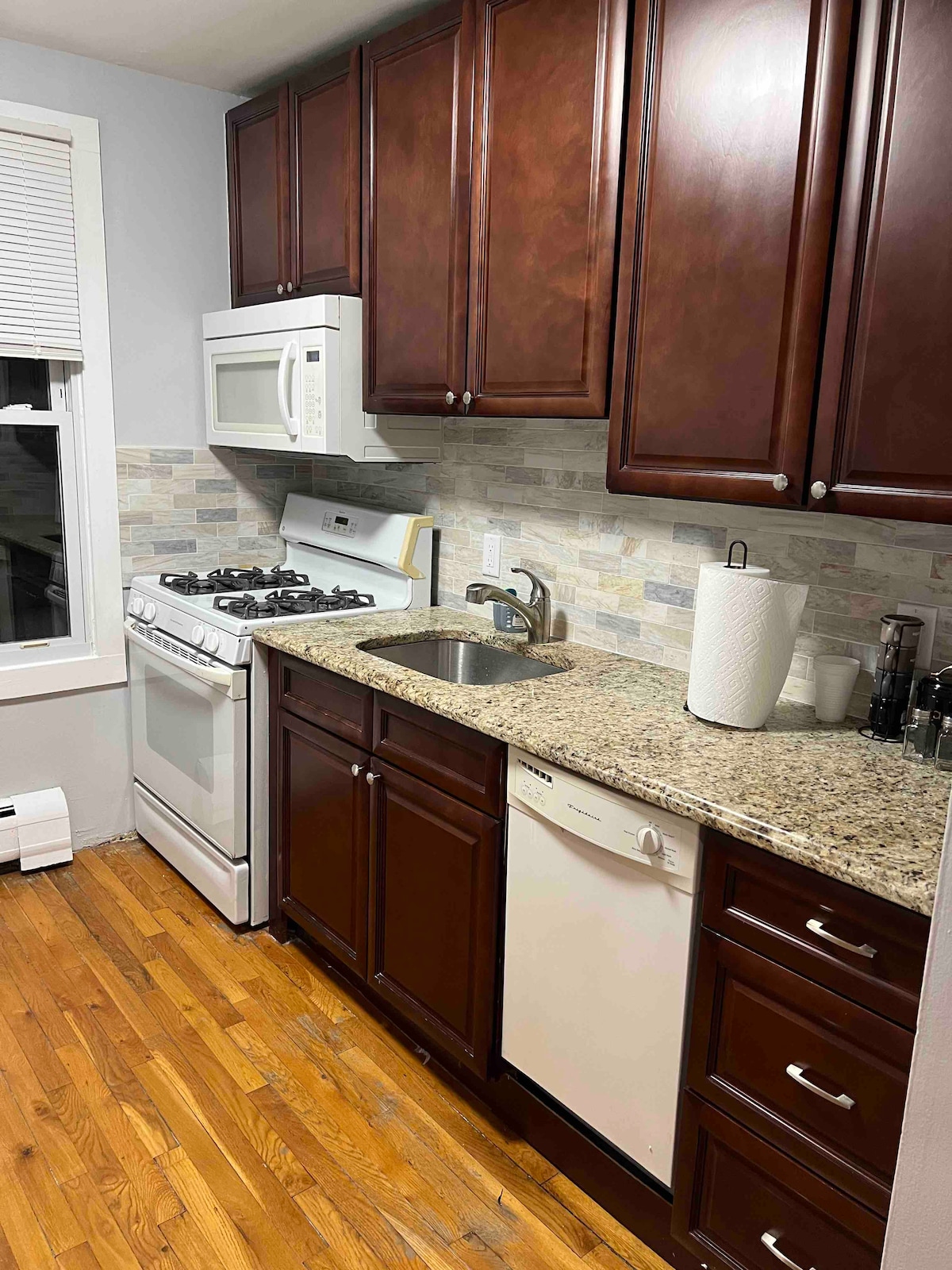 A compact kitchen area features dark wooden cabinets and light-colored granite countertops. Essential appliances include a gas stove, microwave, and dishwasher. A window allows natural light to enter, enhancing visibility. The hardwood floor adds warmth to the space.