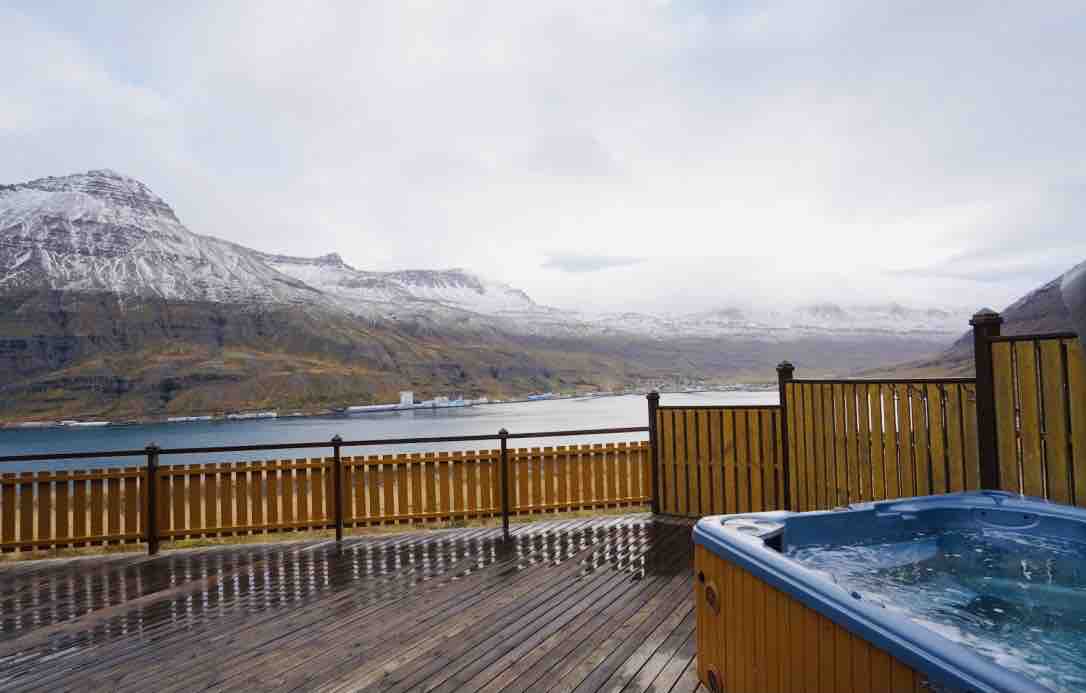 A wooden terrace is shown with a hot tub on one side. The surrounding area features a panoramic view of snow-capped mountains and a calm fjord, enhancing the sense of serenity. Wooden railings encircle the space, adding a touch of privacy.