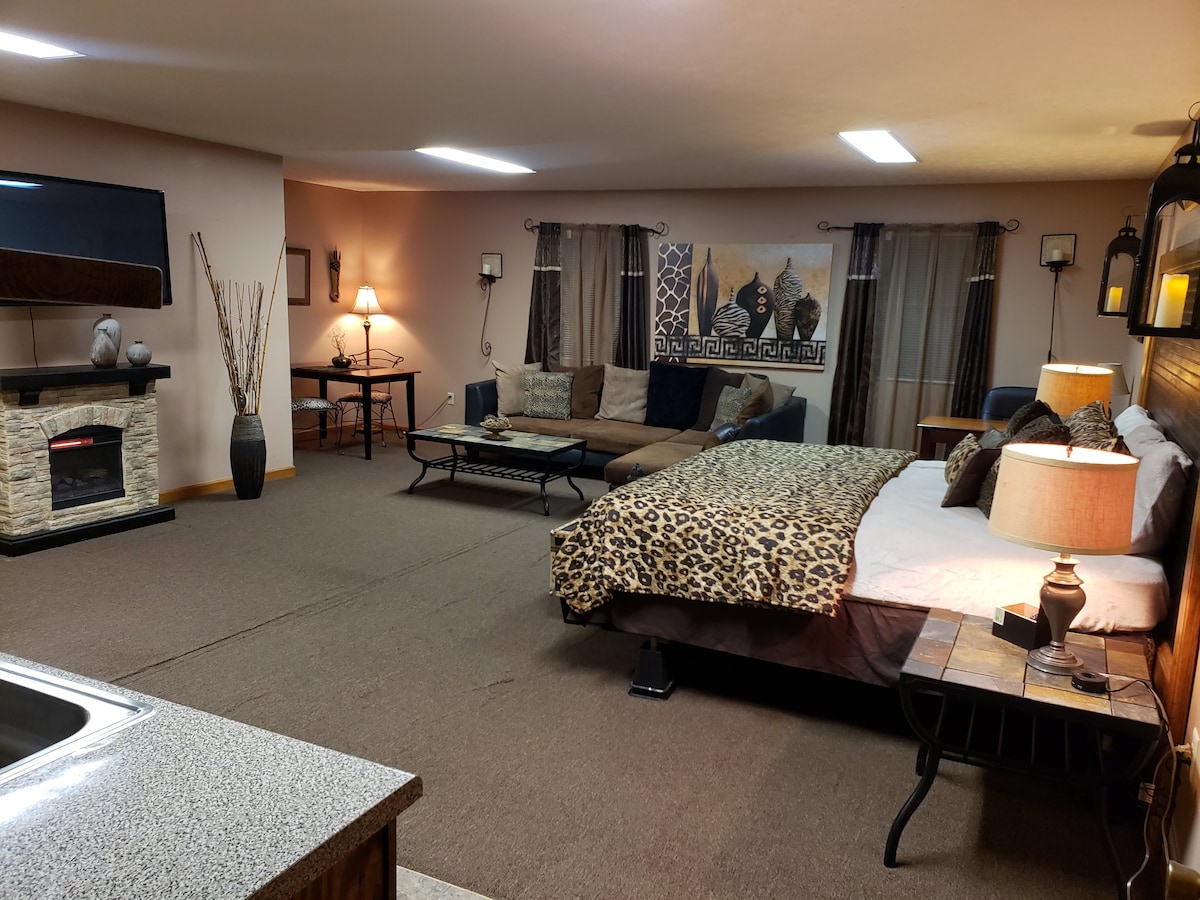 A spacious studio apartment is presented, featuring a King-sized bed with patterned bedding and a cozy electric fireplace. A large TV is mounted on the wall, and an oversized sectional sofa is positioned nearby. Warm lighting is provided by lamps and sconces throughout the room.