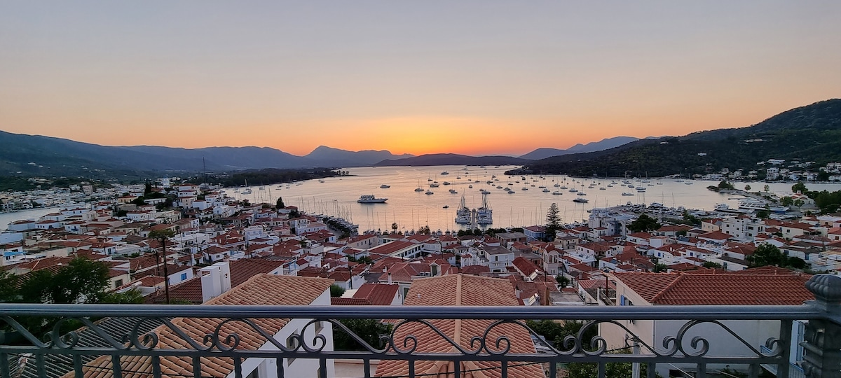 A panoramic view captures the tranquil harbor at sunset, with boats gently anchored in the calm water. The landscape features a blend of traditional white buildings with red roofs, framed by distant mountains and a clear sky transitioning to evening hues.