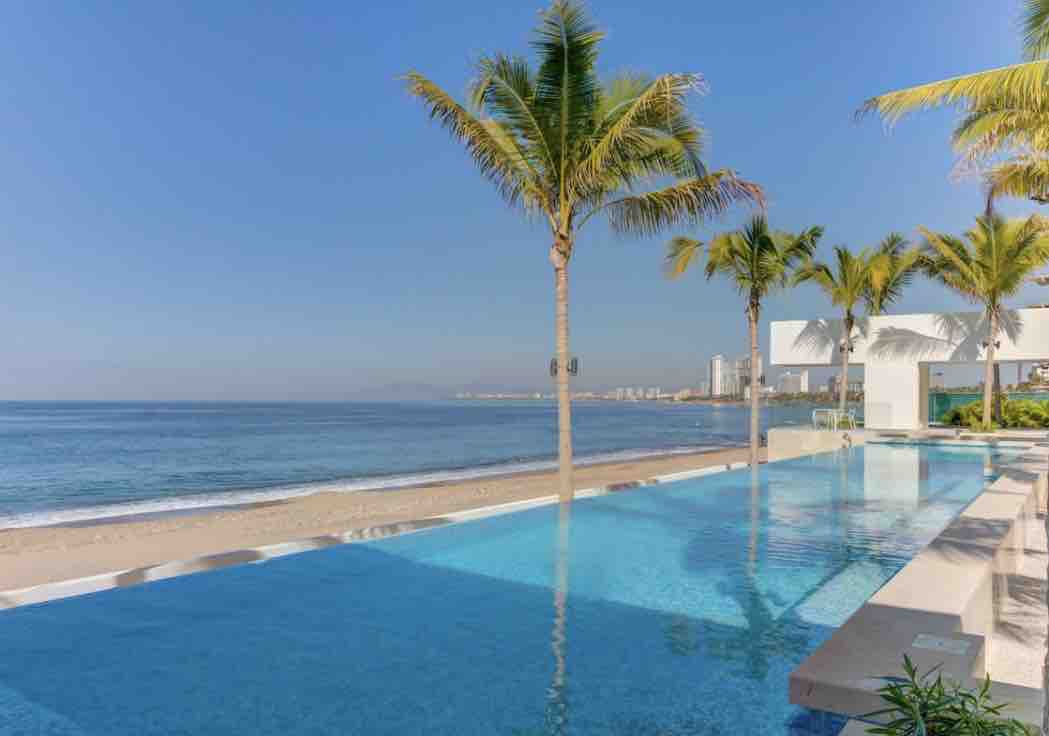 An infinity pool extends towards the horizon, bordered by palm trees. The ocean is visible ahead, with gentle waves lapping against a sandy beach. The backdrop features a clear blue sky, creating a serene seaside environment.