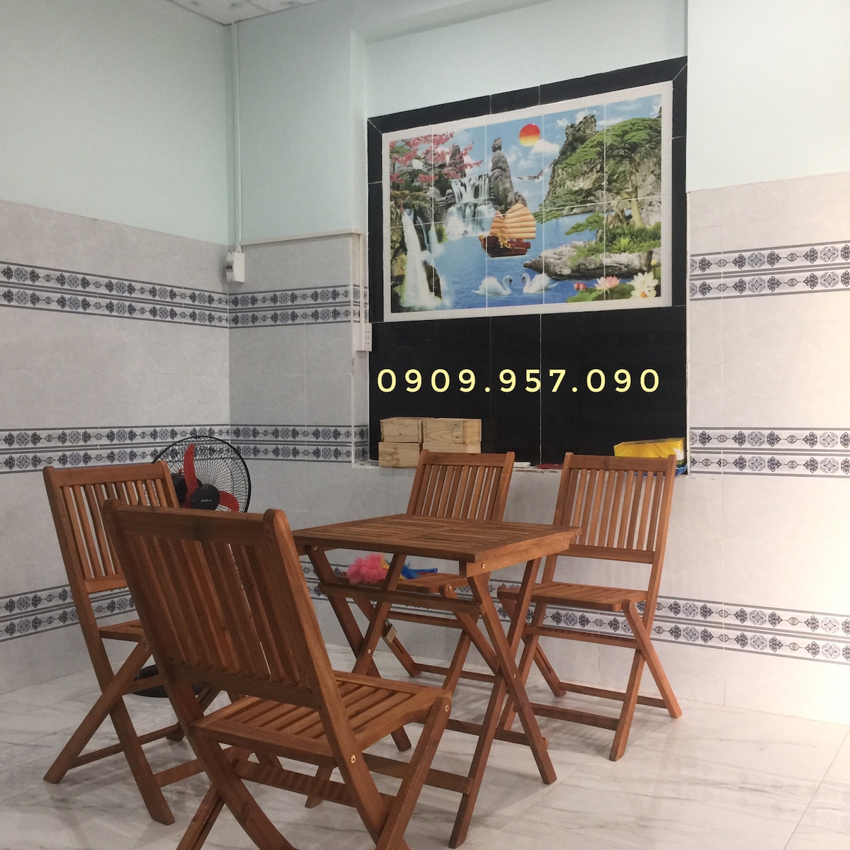 A dining area is presented with a wooden table surrounded by four matching chairs. The walls feature tiled accents and a large mural depicting a serene landscape with waterfalls and mountains. An electric fan is placed nearby for comfort.
