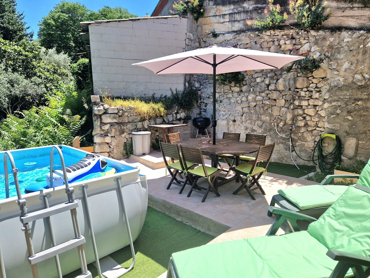 An outdoor space features a private above-ground pool and a wooden dining table surrounded by six chairs, shaded by a large umbrella. Lounge chairs in a light green hue are positioned nearby, against a backdrop of a stone wall adorned with greenery.