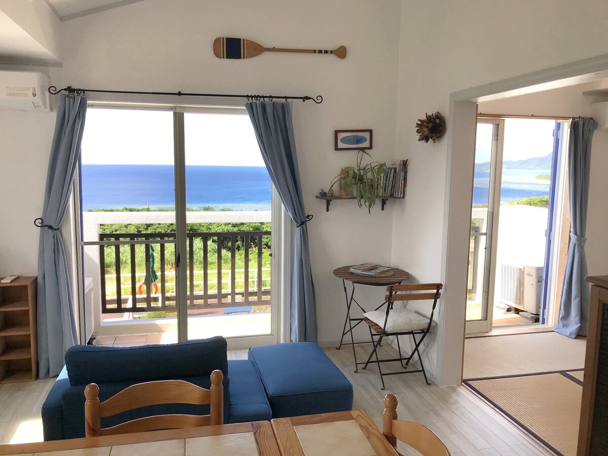 The living area features a blue sofa with wooden dining chairs positioned around a table. Large sliding glass doors open to a balcony with ocean views, while blue curtains frame the windows, allowing natural light to fill the space.