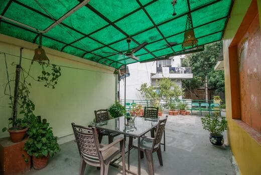 A covered outdoor area features a glass dining table surrounded by four chairs. Greenery is present in pots around the space, with ceiling fans installed overhead. The green canopy provides shade, creating a comfortable environment for dining and relaxation.