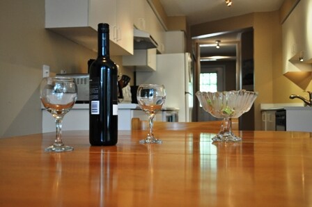 A kitchen countertop is featured with a wooden table displaying two glass goblets and a bottle of red wine, alongside a crystal bowl. The background reveals kitchen appliances and cabinetry, creating a functional and inviting cooking space.