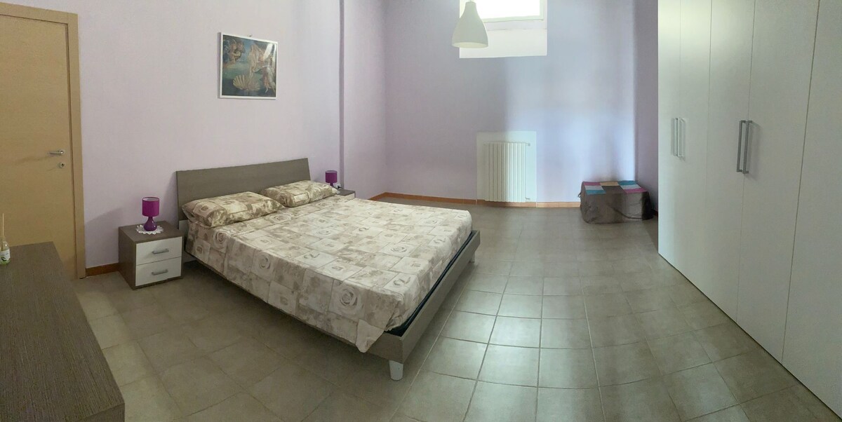 A spacious bedroom features light purple walls and tiled flooring. A large bed with a patterned duvet is positioned centrally. Nightstands with lamps are placed on either side. A wardrobe with multiple doors and a window allowing natural light are also visible.