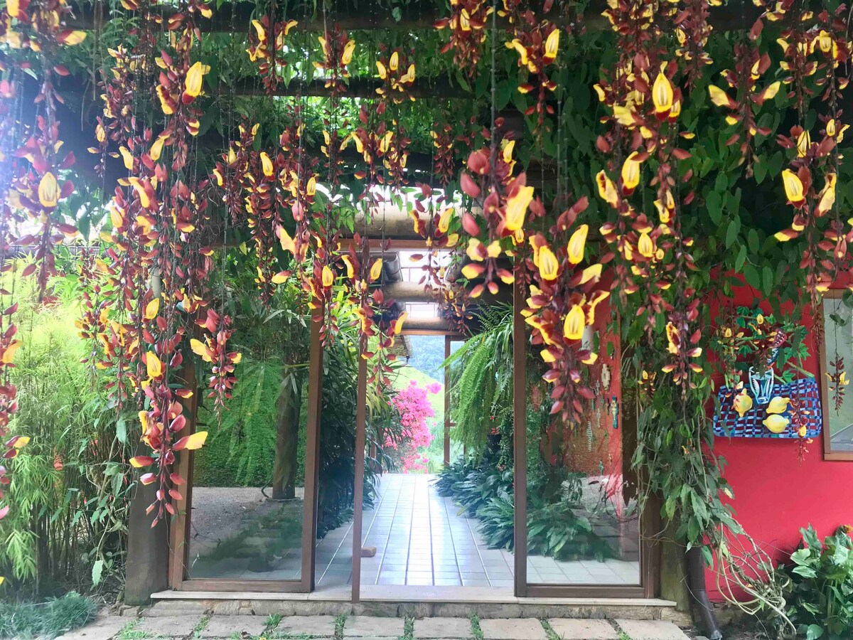 A vibrant entrance is framed by abundant foliage and colorful flowers. The pathway leads through a wooden archway, with glass doors inviting guests into the space. Bright green plants and blooming hues create an inviting atmosphere that connects the indoors to the outdoors.