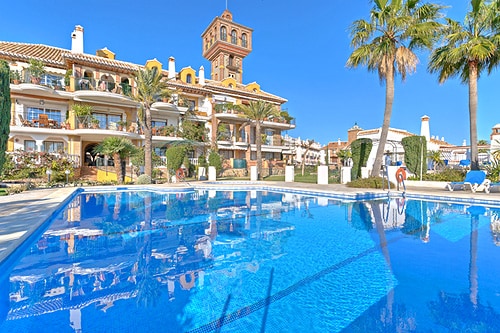 A large swimming pool reflects the clear blue sky, surrounded by well-maintained gardens. The complex is framed by buildings featuring Mediterranean architecture, with palm trees adding a tropical touch. Sun loungers are positioned along the poolside for relaxation.