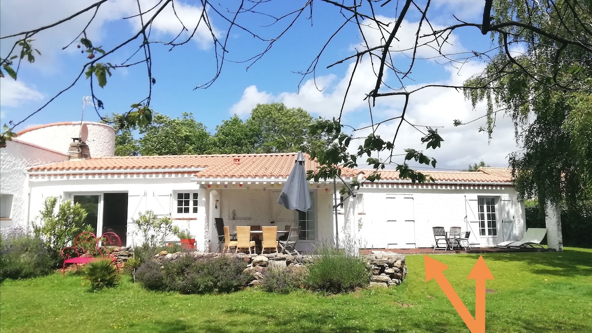 The exterior of the property is shown, featuring a charming white structure surrounded by greenery and flowering plants. A patio area with dining furniture and reclining chairs is visible, set against a backdrop of a clear blue sky and scattered clouds.