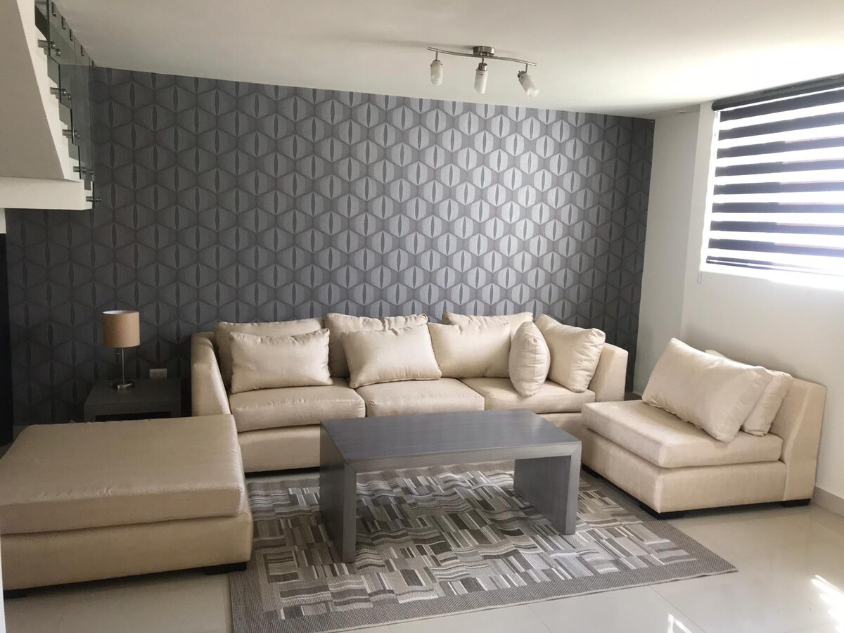 A modern living area features a spacious light-colored sectional sofa, complemented by a stylish coffee table and a decorative lamp. A textured gray wall adds depth, while natural light is filtered through large windows with striped shades, creating a welcoming environment.