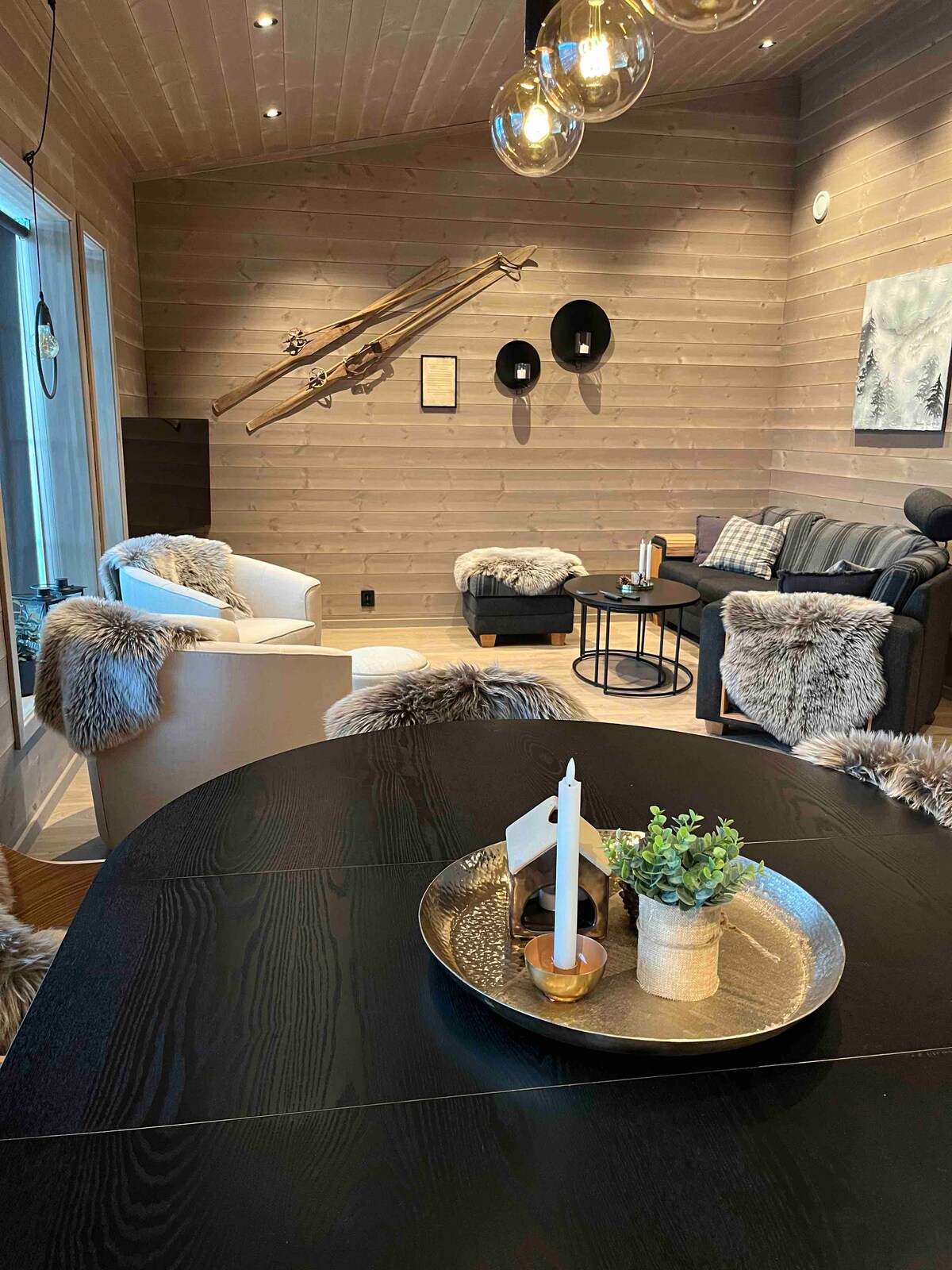 A cozy living area features plush seating covered in soft textures, paired with a sleek coffee table. Natural wood walls create a warm feel, while decorative items enhance the space's charm. A dining table is set with simple accents, providing a welcoming atmosphere.