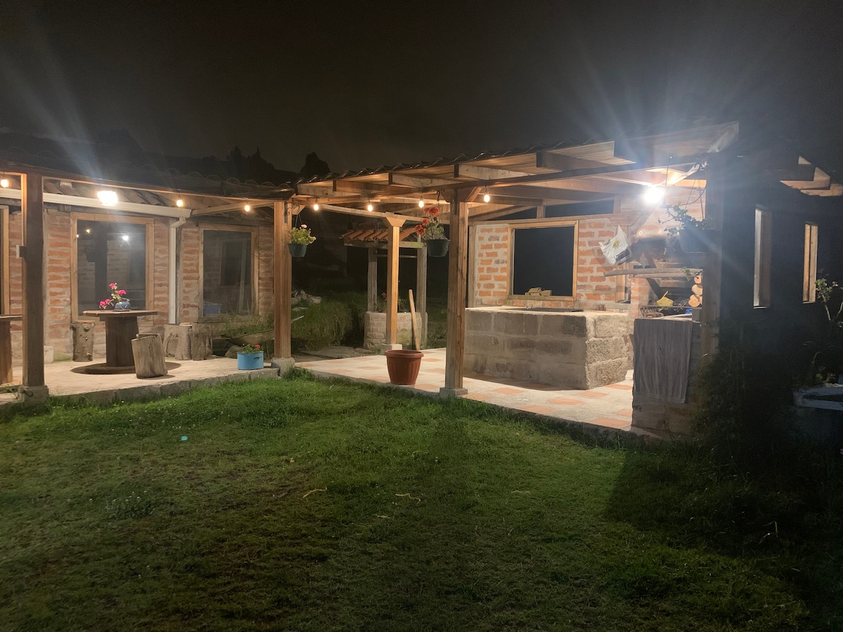 A welcoming outdoor area is illuminated by soft lights, highlighting the wooden beams overhead. Potted plants and flowers are arranged around the space, while a stone structure is partially visible in the background. The grassy area adds to the tranquil atmosphere.