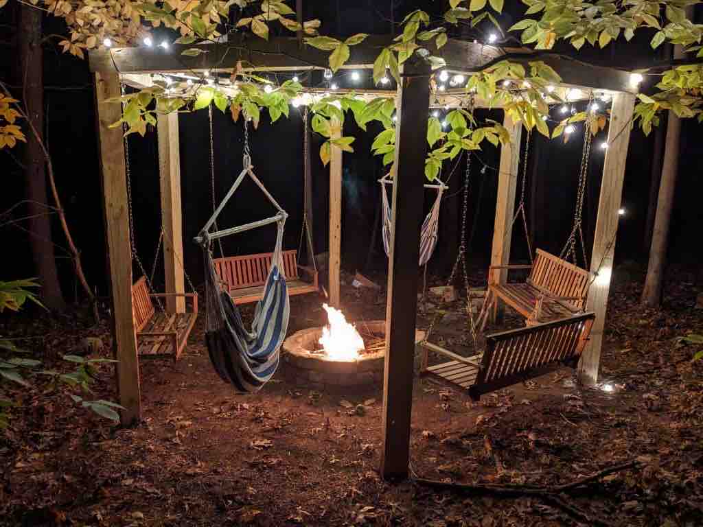 A cozy fire pit area is illuminated by string lights, surrounded by wooden swing chairs and a central stone fire ring. The setting is encased in trees, providing a secluded atmosphere, with fallen leaves on the ground enhancing the natural environment.
