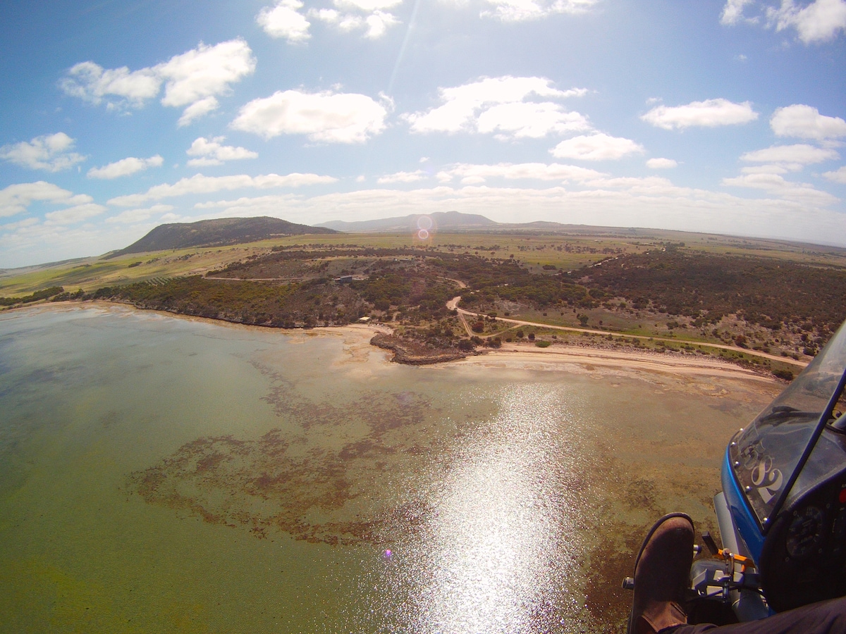 An aerial view captures the shoreline bordered by calm waters, with gentle waves reflecting sunlight. The landscape showcases a mix of land and water, with rolling hills in the background and a winding path visible along the coast.