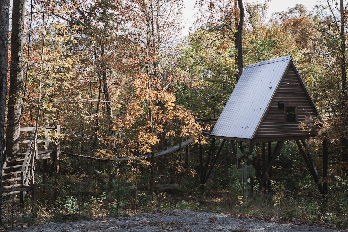 An A-frame treehouse is nestled among vibrant autumn foliage, surrounded by tall trees. A wooden swinging rope bridge connects the treehouse to the ground, creating a whimsical access point. The structure features a sloped metal roof and natural wood siding, harmonizing with the rural landscape.