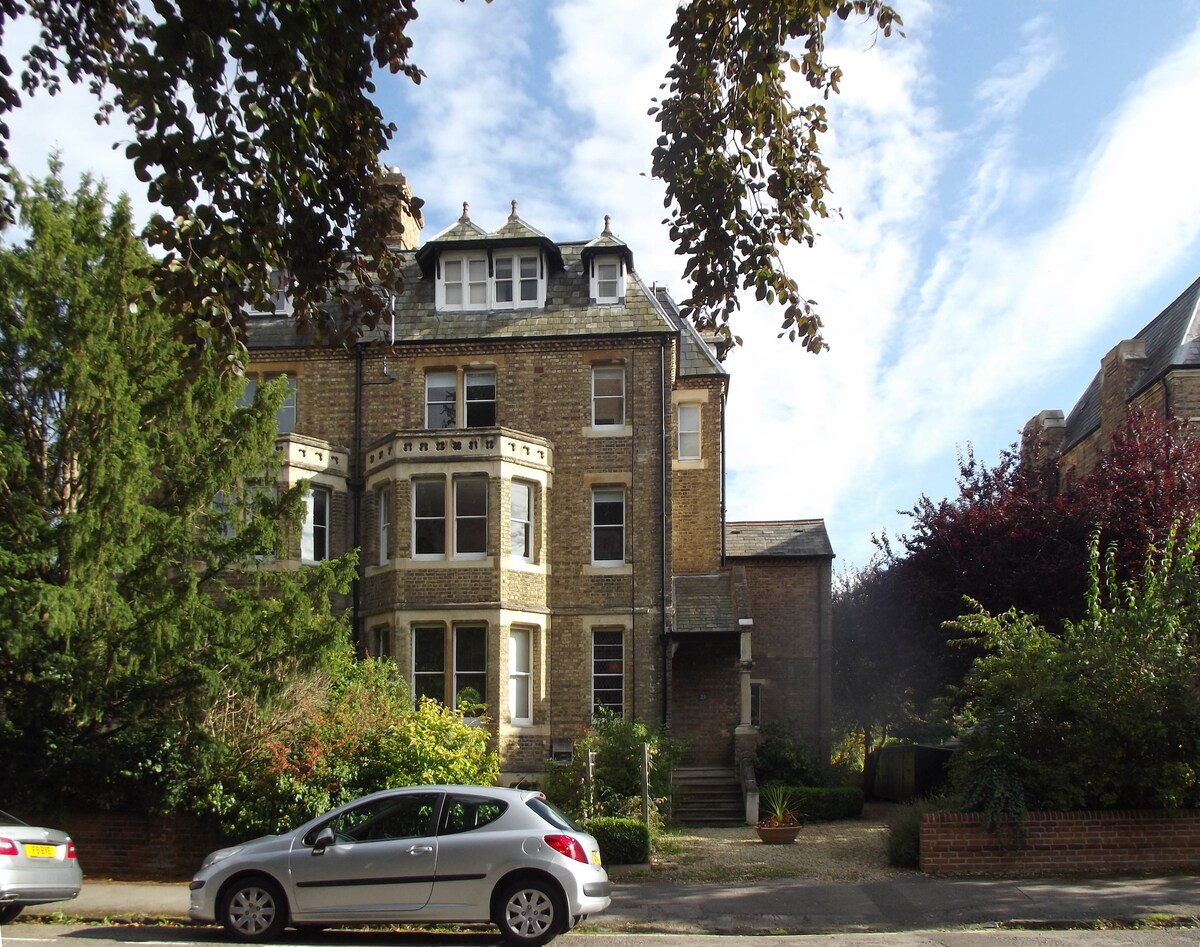 A Victorian house is presented, featuring a distinct stone facade and multiple bay windows. The well-maintained garden is visible, surrounded by lush greenery. A parked car sits along the tree-lined street, enhancing the suburban atmosphere of the conservation area.
