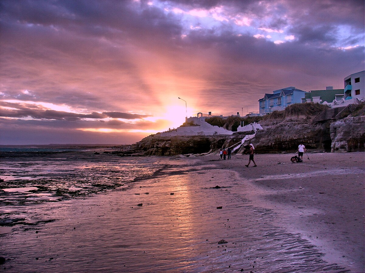 The image features a beach at sunset, with vibrant hues of purple and orange illuminating the sky. Shadows of people walking along the wet sand are visible in the foreground, while calm waves reflect the setting sun. Buildings are seen in the background, adding context to the coastal environment.