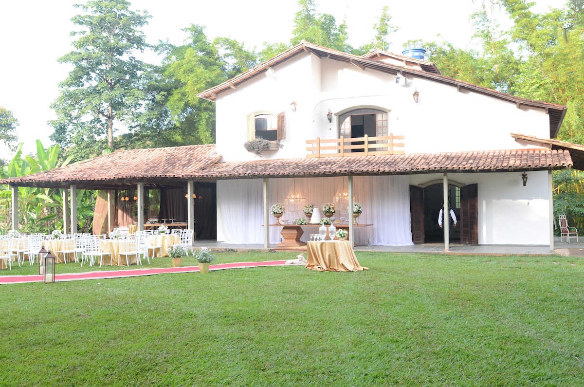 The exterior of the spacious house features a traditional roof and large windows. A well-maintained lawn is present, with elegantly set tables and chairs for outdoor gatherings. Decorative elements add a festive touch, complemented by surrounding lush greenery.