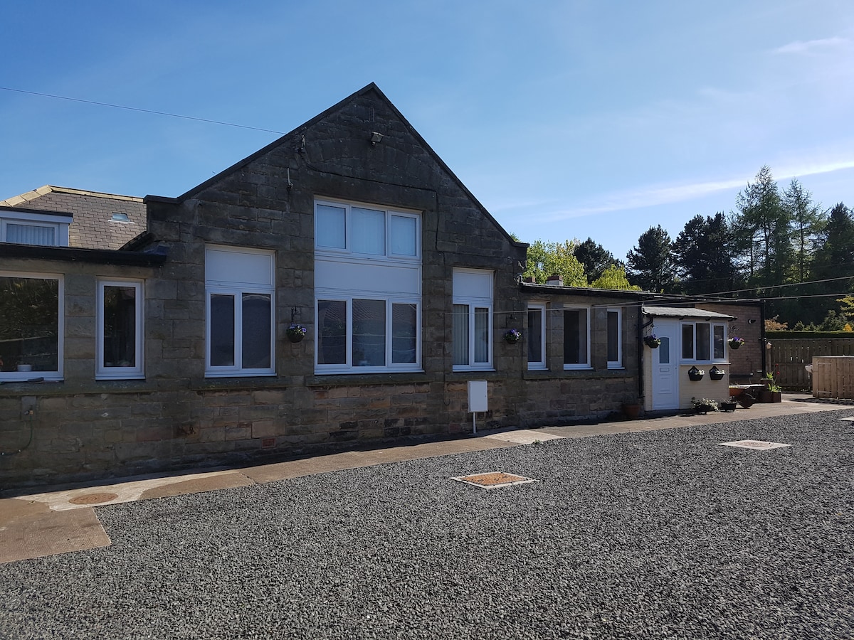 The exterior of the property displays a stone-clad facade, featuring large windows that allow natural light to fill the space. A gravel driveway leads to the entrance, which includes a door framed by flower pots. Surrounding greenery enhances the tranquil setting.