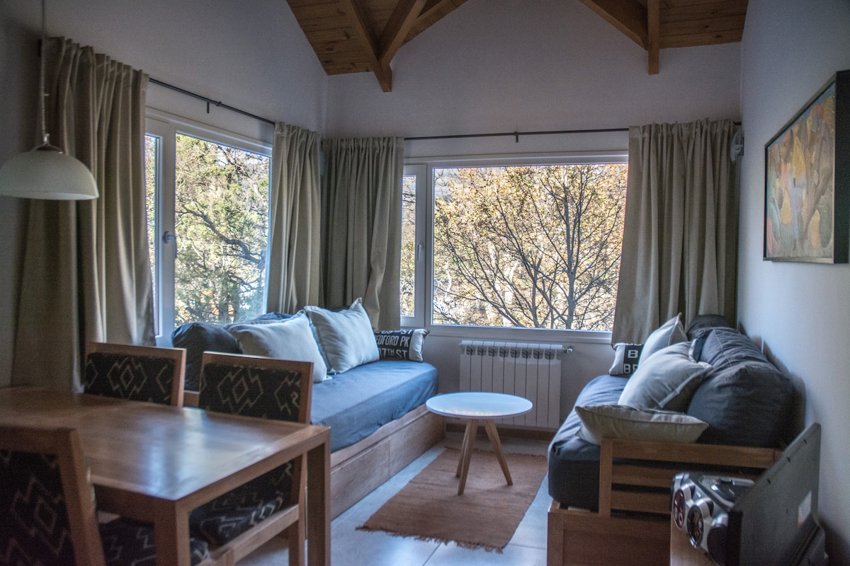 A well-lit living area features large windows offering views of the surrounding trees. Comfortable seating is provided on a sofa and a daybed. A simple table with chairs is positioned nearby, and warmth is accentuated by wooden details and a soft area rug.