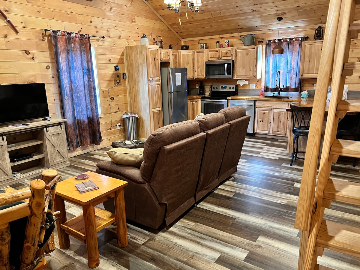 A cozy living area is furnished with a comfortable brown couch and an end table. Light wood walls and flooring create a warm ambiance. The kitchen features wood cabinetry and modern appliances, with a dining area visible and large windows dressed in dark curtains, allowing for natural light.