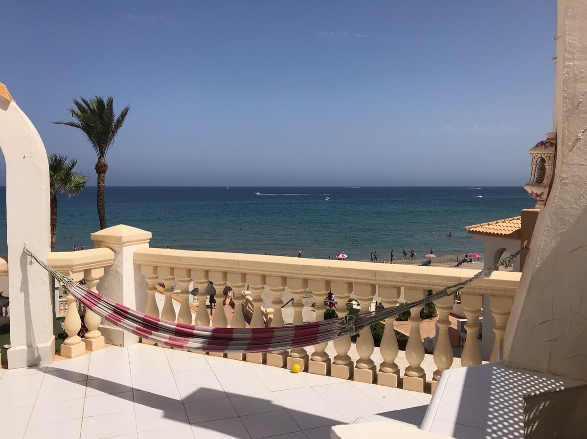 A balcony space is depicted with a colorful hammock suspended between two pillars. The view extends to a serene beach, where a gentle ocean meets the sunny sky. Palm trees frame the scene, creating a sense of relaxation and coastal atmosphere.